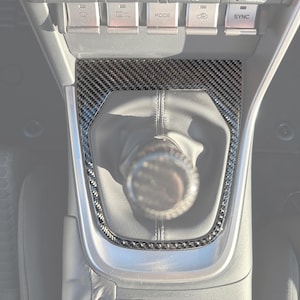 Può includere: Console interna dell'auto con finiture in fibra di carbonio attorno al cambio e al pannello di controllo. La console presenta pulsanti per il controllo del clima e altre funzioni del veicolo. Il pomello del cambio ha un design in fibra di carbonio.