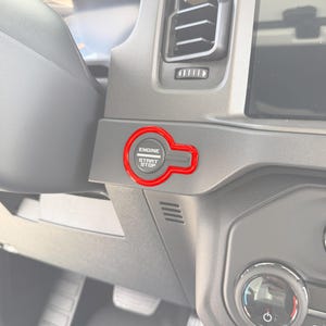 Może przedstawiać: Czerwona osłona przycisku start/stop silnika z białym napisem "ENGINE START STOP". Osłona ma jasnoczerwony kolor i jest zamontowana na szarej desce rozdzielczej. Obraz to zbliżenie wnętrza samochodu.