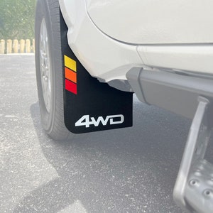 May include: Black mud flaps with a white "4WD" logo and a red, orange and yellow stripe design. The mud flaps are attached to a white vehicle.