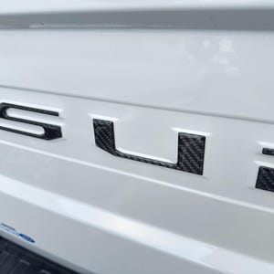 Puede incluir: Un vehículo blanco con un emblema "SURF" de fibra de carbono negra en la parte trasera.