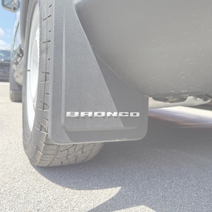 May include: A close-up of a black mud flap on a vehicle, with the silver word "BRONCO" across the bottom. The tyre and wheel are visible, along with a portion of the vehicle's body.