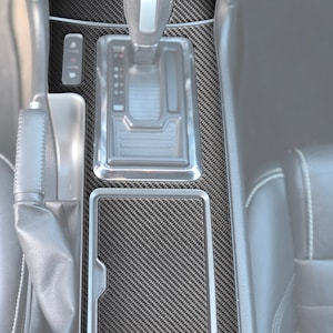 May include: A black carbon fiber center console trim kit for a car. The kit includes a piece for the area around the gear shifter and a piece for the area below the gear shifter.