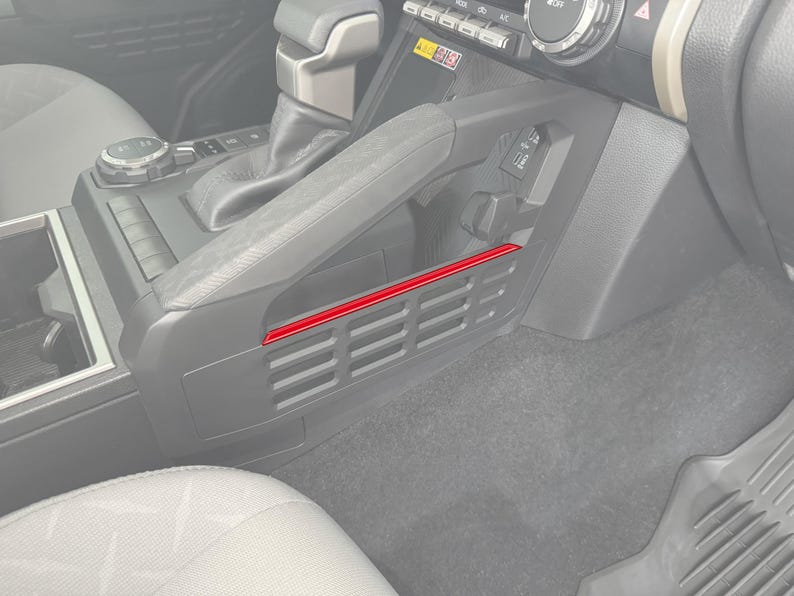 May include: A gray car interior with a black plastic storage compartment with a red trim. The compartment has a series of small rectangular openings.