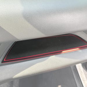 May include: A black rectangular car interior accessory with a red trim. The accessory is set into a gray dashboard compartment. The image is a close-up shot of the car interior.