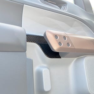 May include: Interior car door panel featuring a rose gold armrest with four circular buttons and a textured surface. A black carbon fiber accent is visible, along with a white speaker and other interior components.
