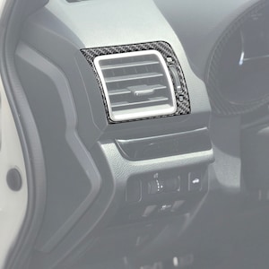 May include: A car dashboard air vent with a carbon fiber trim. The vent is rectangular with horizontal slats and a silver frame. The carbon fiber trim has a black and gray pattern, adding a sporty aesthetic to the vehicle's interior.