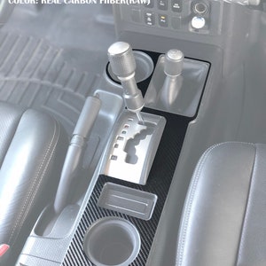 May include: A black carbon fibre centre console trim kit for a car. The kit includes a cup holder, a storage compartment, and a gear shift surround. The trim kit is designed to fit a Toyota FJ Cruiser.