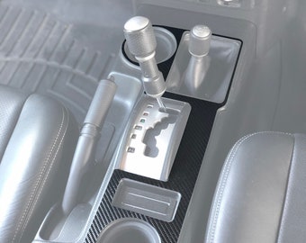Puede incluir: Un kit de moldura de consola central de fibra de carbono negra para un automóvil. El kit incluye un portavasos, un compartimento de almacenamiento y un contorno de palanca de cambios. El kit de moldura está diseñado para adaptarse a un Toyota FJ Cruiser.