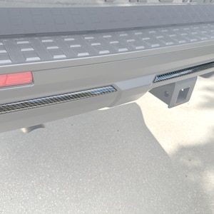 May include: Close-up of a vehicle's rear bumper with carbon fiber trim accents. The bumper is gray with a black trailer hitch receiver. A red reflector is visible on the left side.