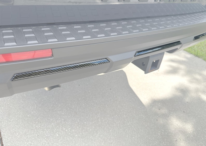May include: Close-up of a vehicle's rear bumper with carbon fiber trim accents. The bumper is gray with a black trailer hitch receiver. A red reflector is visible on the left side.