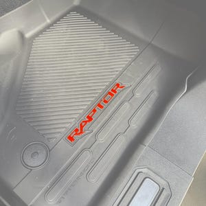 Puede incluir: Alfombrilla negra para el suelo con una superficie texturizada y la palabra "RAPTOR" en letras rojas. La alfombrilla tiene un diseño moldeado, probablemente para un vehículo.