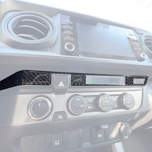 May include: A black carbon fiber dash trim kit for a Toyota Tacoma. The trim kit covers the area above the radio and climate control panel.