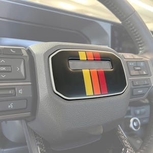 以下が含まれることがあります： 黒とグレーのデザインの車のステアリングホイールのクローズアップ。ステアリングホイールには、赤、黄、オレンジのストライプデザインの黒いパネルがあります。パネルの中央には小さな長方形の開口部があります。
