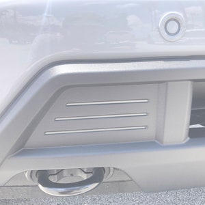 May include: A close-up of a grey vehicle bumper with a black trim. The bumper has three horizontal silver lines and a circular tow hook. A sensor is visible above the bumper.