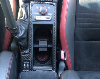 May include: Close-up of a car's interior with a black leather centre console, red stitching, and a carbon fibre trim. The console features a gear shifter, cup holders, and a control panel with the text "SI-DRIVE" and "Sport" buttons.