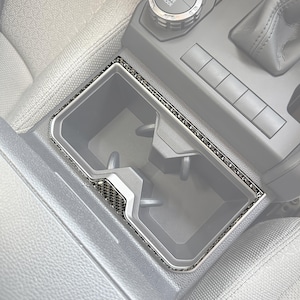 以下が含まれることがあります： カーボンファイバーのトリムが付いた車のセンターコンソールのカップホルダー。カップホルダーは長方形で、2つの円形の開口部があります。トリムはシルバーの縁取りと、黒とシルバーのカーボンファイバーパターンです。