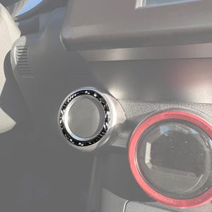 Può includere: Primo piano dell'interno di un'auto, con un pomello di controllo argentato e nero. La manopola circolare ha un anello nero strutturato. Sono visibili anche un indicatore con bordo rosso e un pulsante "AUTO".