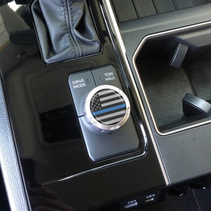 May include: A close-up of a car's interior showing a black and silver knob with a thin blue line American flag design. The knob is labelled "Drive Mode" and "Tow/Haul".