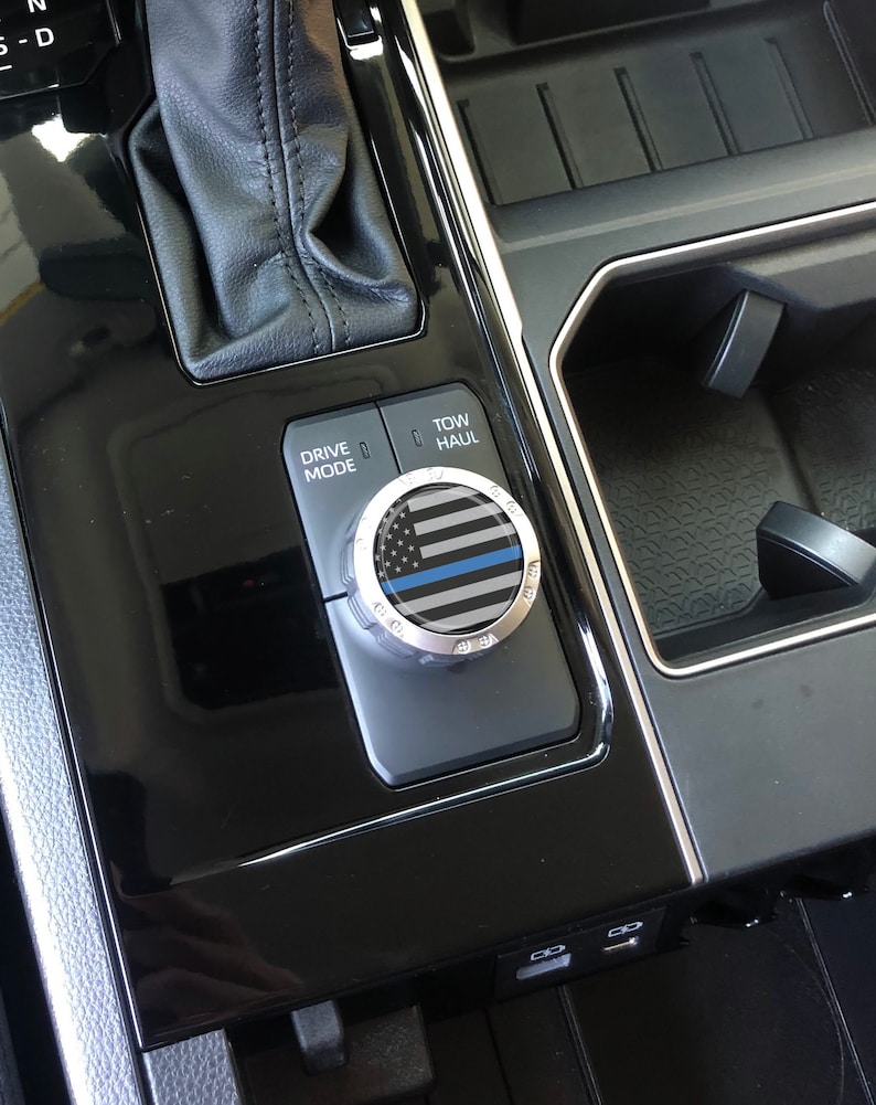 May include: A close-up of a car's interior showing a black and silver knob with a thin blue line American flag design. The knob is labelled "Drive Mode" and "Tow/Haul".