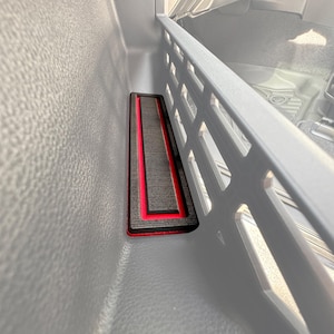 Pode incluir: Inserção retangular da consola do carro com acabamento preto e vermelho. A inserção foi concebida para caber na área da consola de um veículo. O interior da consola é cinzento.