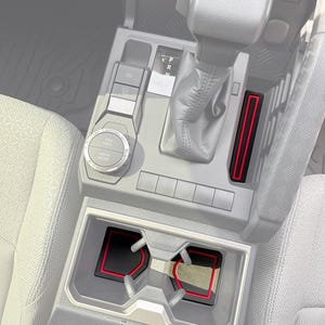 Könnte beinhalten: Set mit Innenzubehör für Autos. Das Bild zeigt die Mittelkonsole eines Autos mit schwarzen und roten Akzentteilen. Das Set enthält Becherhaltereinsätze und eine Konsolen-Tablettauskleidung, alle mit rotem Rand. Der Schalthebel und das Bedienfeld des Autos sind ebenfalls sichtbar.