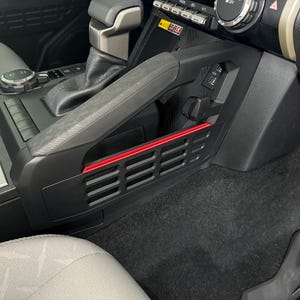 May include: A black car interior with a red trim on the side of the center console. The console has a black plastic vent with a grid pattern.