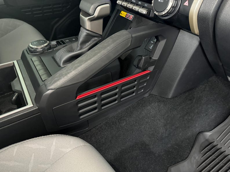 May include: A black car interior with a red trim on the side of the center console. The console has a black plastic vent with a grid pattern.