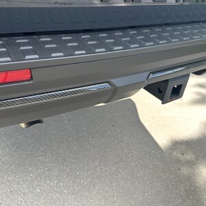 May include: Close-up of a vehicle's rear bumper, featuring a black trailer hitch receiver. The bumper is gray with a textured black top section and carbon fiber trim. A red taillight is visible on the left.