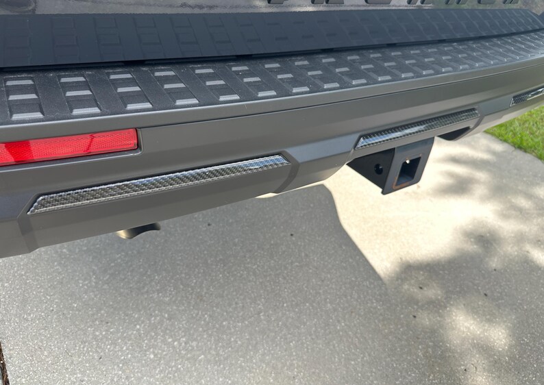 May include: Close-up of a vehicle's rear bumper, featuring a black trailer hitch receiver. The bumper is gray with a textured black top section and carbon fiber trim. A red taillight is visible on the left.
