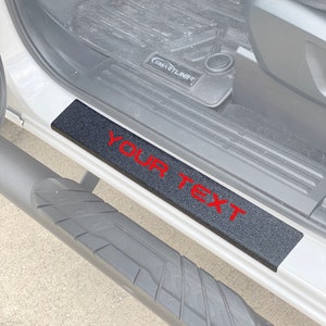 Puede incluir: Peldaño de camión negro con una inscripción roja "YOUR TEXT" en una superficie negra texturizada. El peldaño está fijado al lado de un camión blanco.