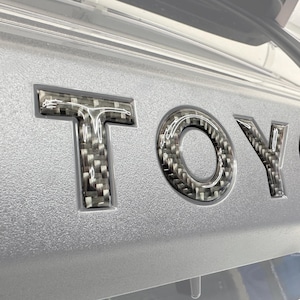 Puede incluir: Primer plano de un panel de coche plateado con la palabra "TOYO" en relieve en un patrón de fibra de carbono. Las letras están en relieve y tienen un acabado brillante, contrastando con el fondo plateado texturizado.
