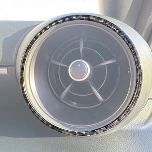 May include: Close-up of a car's air vent with a carbon fiber trim ring. The vent is circular with a central knob and five blades. The surrounding dashboard is gray.