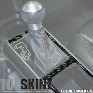 May include: A close-up of a car's interior with a carbon fiber trim kit installed around the gear shifter. The trim kit is a dark gray color with a subtle carbon fiber pattern. The text "COLOR: DOMED CARBON FIBER" is visible at the bottom of the image.