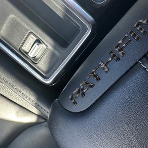 May include: Close-up of a black leather car seat with the word "PATHFINDER" embossed in silver lettering. A silver control panel with a button labeled "D-MODE" is visible in the background.