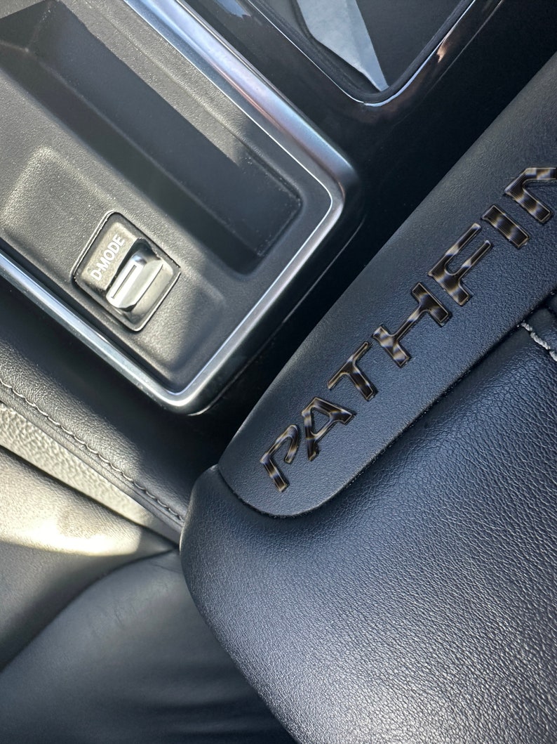 May include: Close-up of a black leather car seat with the word "PATHFINDER" embossed in silver lettering. A silver control panel with a button labeled "D-MODE" is visible in the background.