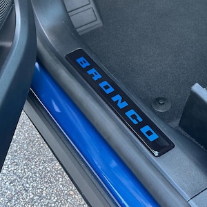 Op de afbeelding: Een zwarte dorpelplaat met het woord "BRONCO" in blauwe letters. De plaat is geïnstalleerd in een voertuig, met een blauw interieurpaneel zichtbaar. De vloer is bedekt met zwart tapijt.