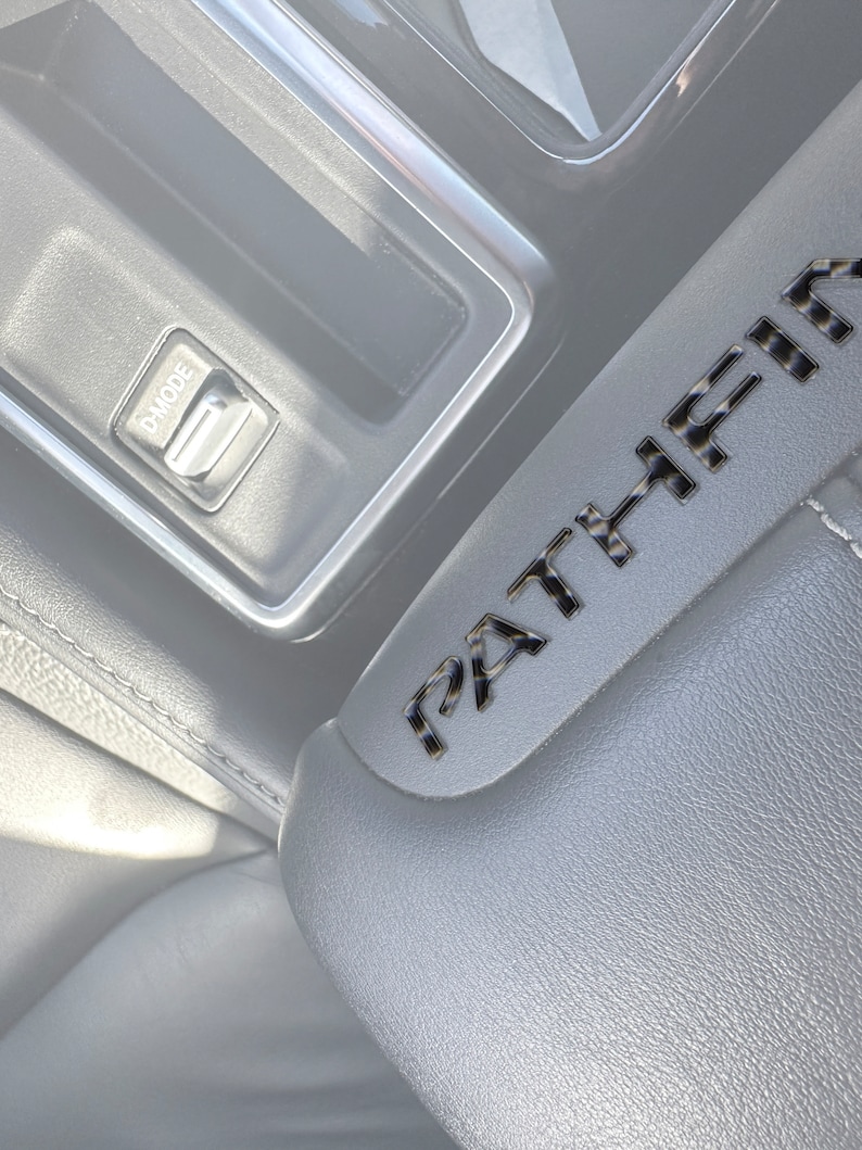 May include: Close-up of a gray leather car seat with a black carbon fiber "PATHFINDER" emblem. A silver button labeled "P-MODE" is visible in the background.