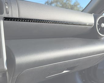 May include: Interior car dashboard with a black trim piece. The dashboard is dark gray with a textured surface. The trim piece is black with a woven pattern. The image is taken on a sunny day.