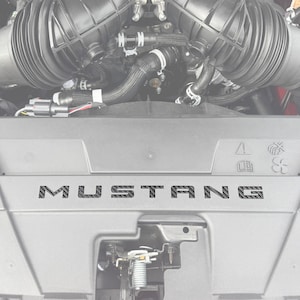 May include: A close-up of a car engine with a black carbon fiber "MUSTANG" emblem on the engine cover.
