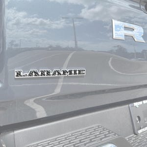 Puede incluir: Primer plano de la parte trasera de un vehículo gris con el emblema cromado "R" y un emblema "LARAMIE". El emblema "LARAMIE" es un rectángulo plateado con texto negro. La superficie del vehículo refleja el cielo y el entorno.