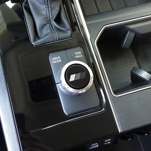 May include: A black car interior with a silver drive mode knob with the text "Drive Mode" and "Tow/Haul". The knob has a black and grey logo in the centre.