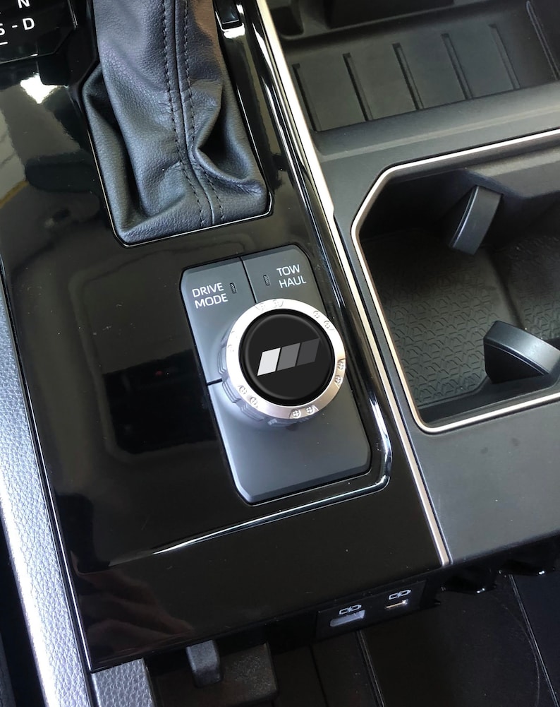 May include: A black car interior with a silver drive mode knob with the text "Drive Mode" and "Tow/Haul". The knob has a black and grey logo in the centre.