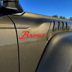May include: Close-up of a bronze-colored vehicle's front quarter panel. The word "Bronco" is displayed in red script. Features include a series of vertical vents and a black tire.