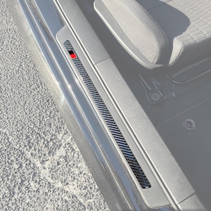 May include: A carbon fiber textured door sill protector strip installed on the interior frame of a vehicle. The long, narrow strip features a black and red rectangular emblem near the front edge, contrasting against the dark car interior.