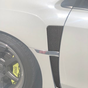 Puede incluir: Un coche blanco con una ventilación de guardabarros de fibra de carbono. La ventilación es negra con un patrón de tejido texturizado. El coche tiene un logotipo "STI" rojo en el lateral.