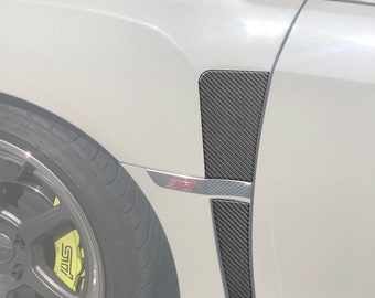 May include: A white car with a carbon fiber fender vent. The vent is black with a textured weave pattern. The car has a red "STI" logo on the side.
