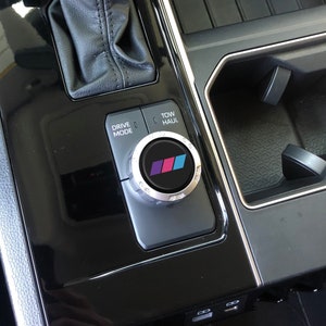 May include: A black car interior with a silver drive mode knob featuring a colourful BMW logo. The knob is labelled "Drive Mode" and "Tow/Haul".