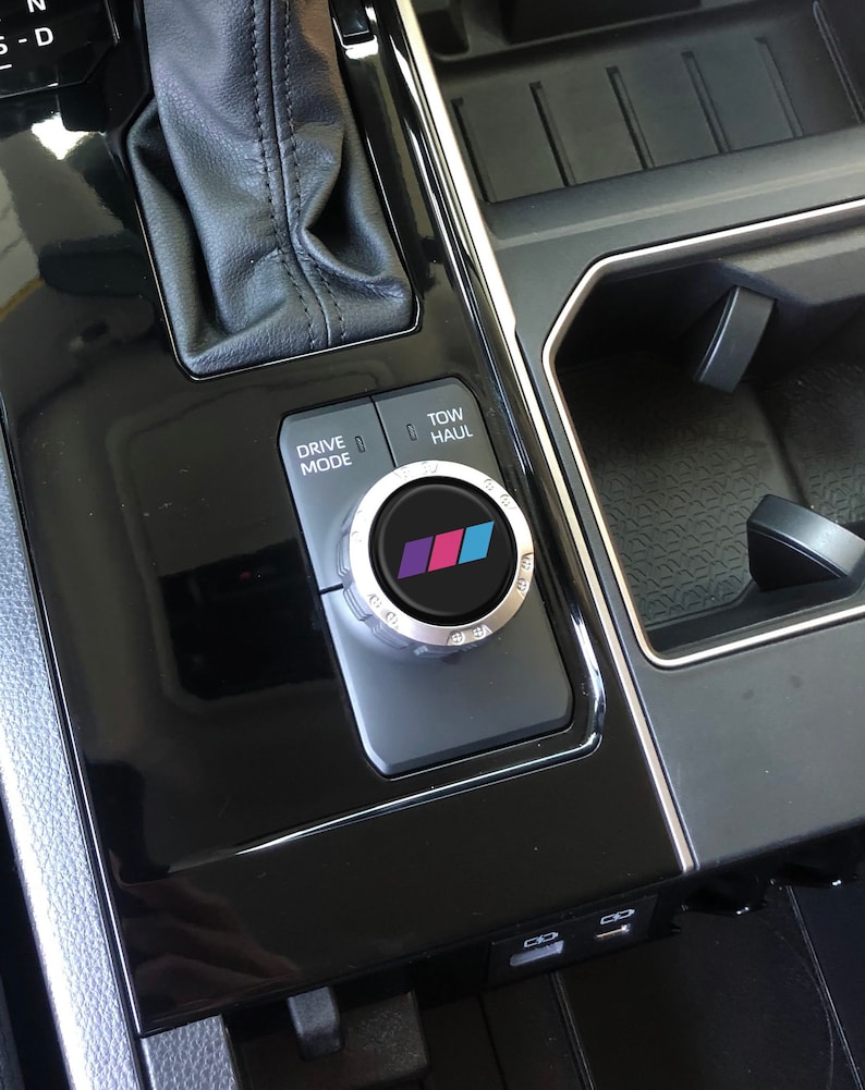 May include: A black car interior with a silver drive mode knob featuring a colourful BMW logo. The knob is labelled "Drive Mode" and "Tow/Haul".