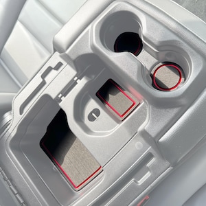 Könnte beinhalten: Eine hellgraue Autokonsole mit schwarzen und grauen Einsätzen und roter Zierleiste. Die Konsole verfügt über zwei Getränkehalter, ein Staufach und einen kleinen rechteckigen Bereich mit rotem Rand.