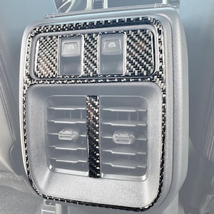 Peut inclure: Garniture intérieure de voiture noire et argentée avec un motif en fibre de carbone. La garniture comprend des boutons de commande de fenêtre et des bouches d'aération. Le bas de la garniture porte l'inscription "AC 110V 400W MAX".